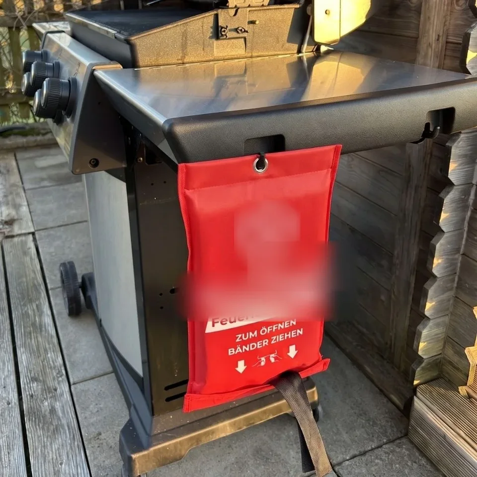 Löschdecke hängt griffbereit an einem Gasgrill auf der Terrasse. Logo unkenntlich gemacht.